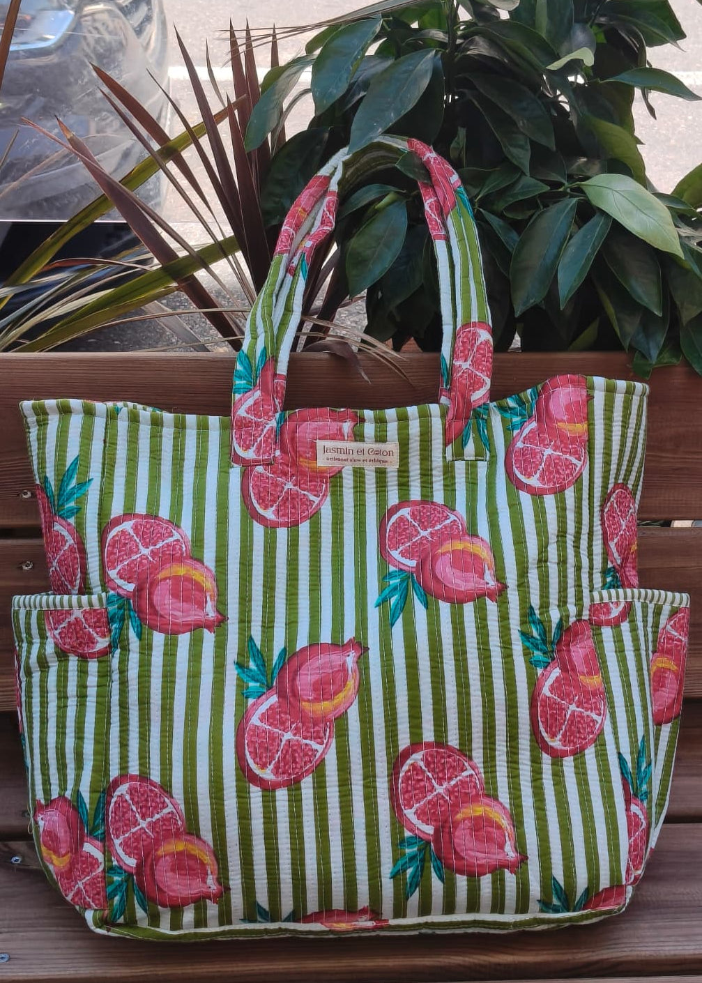 Green striped tote bag with pink fruit pattern on a wooden surface with plants in the background